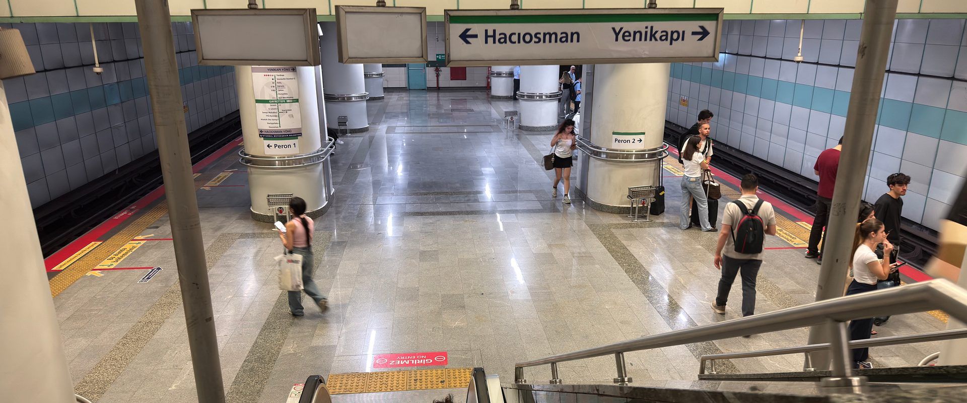 View down an escalator onto an M2 metro platform concourse with Peron 1/2 signs and a few waiting passengers.
